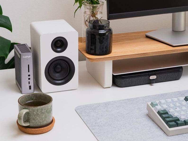 May include: A gray CalDigit docking station, a white speaker with a black driver, a green speckled ceramic mug on a round cork coaster, and a mechanical keyboard with white and green keys arranged on a desk with a monitor stand.