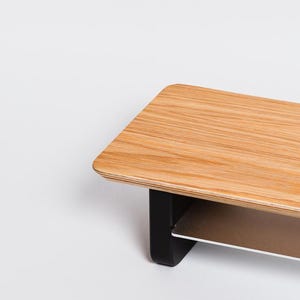 May include: A monitor stand featuring a rectangular light brown wood grain top surface with rounded corners. The stand is supported by two solid black vertical legs and includes a secondary horizontal shelf positioned beneath the main top surface.