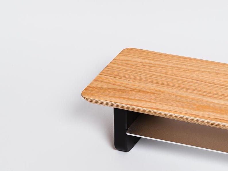 May include: A monitor stand featuring a rectangular light brown wood grain top surface with rounded corners. The stand is supported by two solid black vertical legs and includes a secondary horizontal shelf positioned beneath the main top surface.