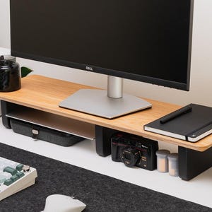 May include: A monitor stand featuring a light wood grain top surface supported by two black legs. The stand provides an elevated platform for a computer monitor, with open storage space underneath for a laptop, a black camera, and a notebook.