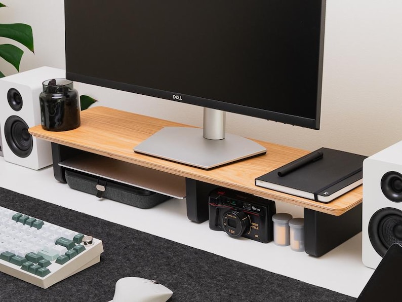 May include: A monitor stand featuring a light wood grain top surface supported by two black legs. The stand provides an elevated platform for a computer monitor, with open storage space underneath for a laptop, a black camera, and a notebook.