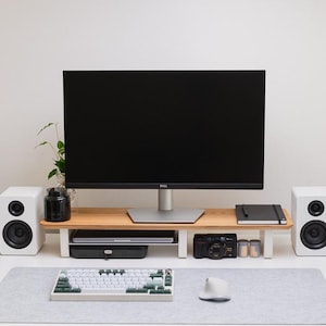 May include: A wooden monitor stand with white legs sits on a desk, supporting a black Dell monitor. Below the stand are a laptop and a camera. Flanking the stand are two white speakers. A mechanical keyboard and a white mouse rest on a gray desk mat.