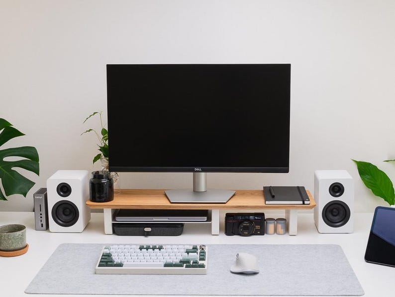 May include: A wooden monitor stand with white legs sits on a desk, supporting a black Dell monitor. Below the stand are a laptop and a camera. Flanking the stand are two white speakers. A mechanical keyboard and a white mouse rest on a gray desk mat.