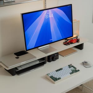 May include: A white monitor stand with black legs holding a computer monitor displaying a blue abstract background with the time 8:44. A silver laptop, a black camera, a red toy race car, a notebook, a white mouse, and a white and green keyboard.