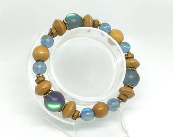 May include: A beaded bracelet featuring a mix of wooden, iridescent, and blue glass beads. The bracelet has a variety of bead shapes and sizes, with gold-colored spacers. The bracelet is displayed on a clear stand.