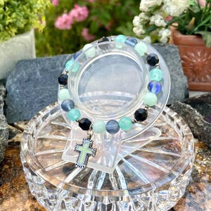 Puede incluir: Una pulsera de cuentas con un colgante de cruz. La pulsera presenta una mezcla de cuentas azules, verdes y negras. El colgante de cruz tiene un centro azul claro y un marco plateado. La pulsera se exhibe en un soporte decorativo transparente.