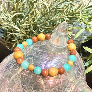 Op de afbeelding: Een armband met ronde kralen in goud, bruin en turquoise. De armband wordt getoond op een decoratief glazen object, met groene bladeren op de achtergrond. De armband is een sieraad.