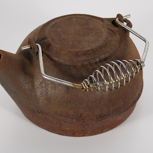 May include: A round, dark brown, rusted cast iron kettle with a short spout and a circular lid. The kettle features a silver-colored metal bail handle with a coiled wire grip attached to two small lugs on the top of the kettle body.