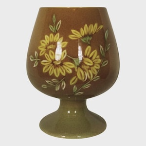 May include: A brown ceramic vase with a wide, rounded top and a pedestal base. The vase is decorated with yellow sunflowers and green leaves. The vase is approximately 6 inches tall.