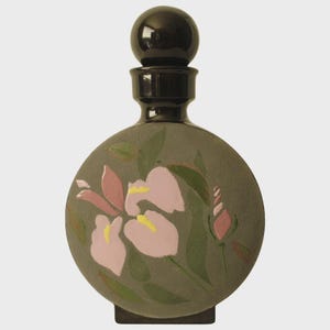 May include: A round, dark gray ceramic bottle with a black stopper. The bottle is decorated with pink and yellow flowers and green leaves. The bottle has a square base.