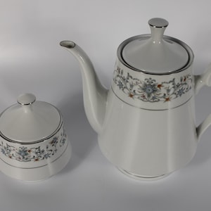 May include: A white porcelain teapot and a matching lidded sugar bowl, both featuring a delicate floral pattern in shades of blue, grey, and orange, accented with thin silver trim along the rims, handles, and the pointed lids of each piece.