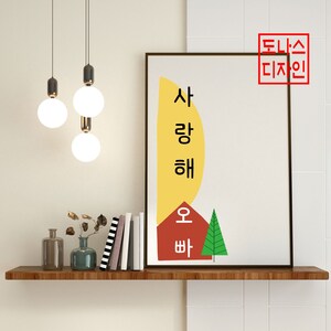 May include: A framed art print with Korean text and a minimalist design. The artwork features a yellow shape, a red house, and a green tree. The text says "I love you, Oppa". The print is displayed on a wooden shelf.