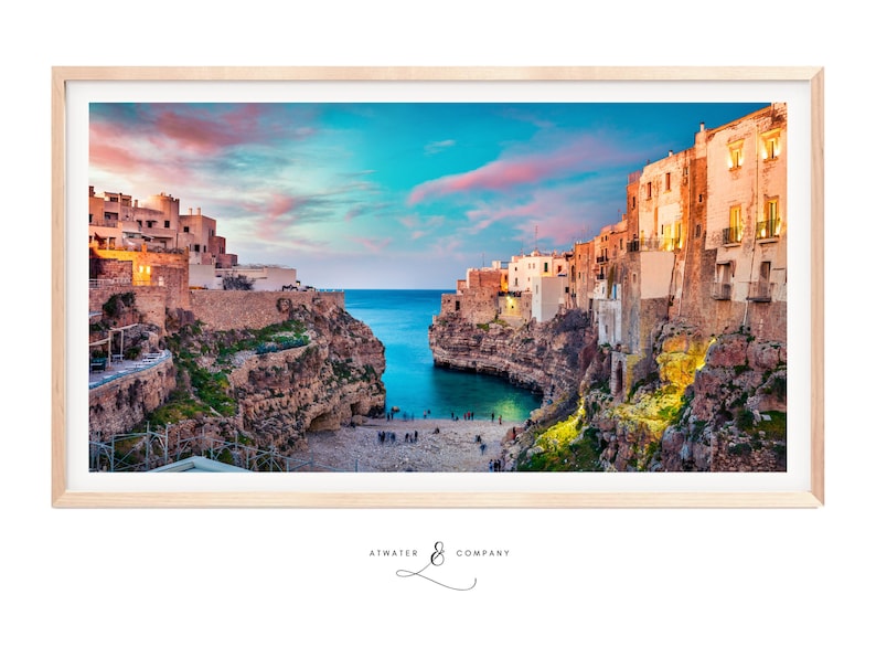 May include: A panoramic photograph of a coastal town at sunset. The town is built on cliffs overlooking a small bay. The sky is a vibrant blue and pink, and the water is a deep blue. The buildings are white and have terracotta roofs. There are people on the beach below the cliffs.