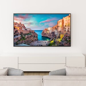 May include: A white couch with two pillows sits in front of a white entertainment center with a flat screen TV displaying a scenic view of a coastal town with buildings on cliffs overlooking a bay.