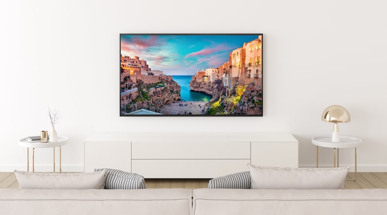 May include: A white couch with two pillows sits in front of a white entertainment center with a flat screen TV displaying a scenic view of a coastal town with buildings on cliffs overlooking a bay.