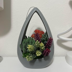 May include: A gray ceramic hanging planter shaped like a teardrop with a variety of artificial succulents and flowers inside.