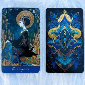 May include: Two rectangular tarot cards featuring deep blue and gold color palettes. The left card depicts a figure in a black gown with a gold halo, wheat stalks, and the text The Empress. The right card shows a symmetrical blue and gold pattern.