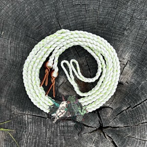 May include: A white and green braided rope with brown leather ends. The rope is coiled on a wooden surface. The text "Unrivaled Tack Co." is visible in the lower center of the image.