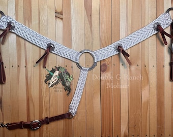 Könnte beinhalten: Ein weißes Mohair-Brustgeschirr mit silbernen Beschlägen und Lederriemen. Das Brustgeschirr ist geflochten und hat einen runden Ring in der Mitte. Der Text "Double C Ranch Mohair" ist auf dem Holzgrund sichtbar.