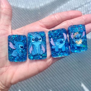 May include: Four rectangular, blue glitter resin pieces featuring the character Stitch from Lilo & Stitch. Each piece has a different pose of Stitch, with varying details and expressions. The pieces are held in a hand.