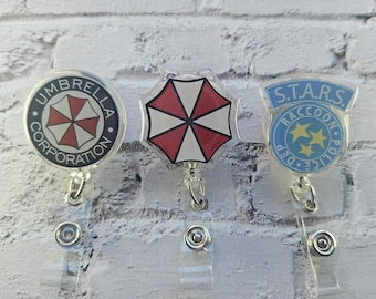 Op de afbeelding: Drie badgehouders met Resident Evil-ontwerpen. Links staat het logo van de Umbrella Corporation. In het midden een rood-witte parapluvorm. Rechts staat het blauwe embleem van het S.T.A.R.S. Raccoon Police Department afgebeeld.