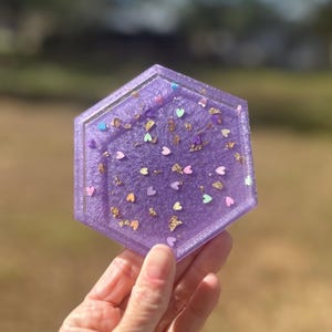 May include: A hexagonal purple resin coaster with a glittery finish. The coaster is decorated with small, colorful hearts and gold flakes.