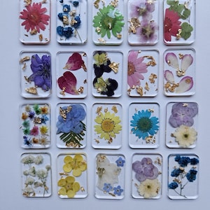 May include: A collection of rectangular resin pieces, each containing a different pressed flower and gold leaf flakes. The flowers vary in color, including red, blue, yellow, and purple. The resin pieces are arranged in a grid pattern.
