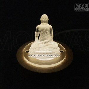 Könnte beinhalten: Eine weiße Glas-Buddha-Statue, die auf einem runden Holzsockel mit einem weißen Licht steht, das von unten scheint. Der Sockel trägt den Text "www.monivas.net".