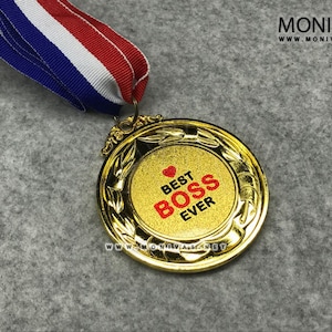 Może przedstawiać: Złoty medal z czerwonym sercem i napisem "Best Boss Ever" na złotym tle. Medal jest przymocowany do niebiesko-biało-czerwonej wstążki.
