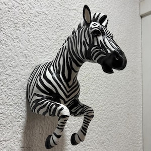 Op de afbeelding: Een zwart-witte zebrahoofd wandsculptuur. De sculptuur is gemonteerd op een getextureerde witte muur. De zebra heeft een gedetailleerd ontwerp met zwarte strepen op een witte achtergrond. De kop van de zebra is opzij gedraaid.