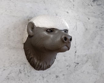 May include: A honey badger head sculpture mounted on a textured wall. The sculpture features a dark gray face and a white top, with detailed features like ears and a nose. The artwork is a unique decorative piece.