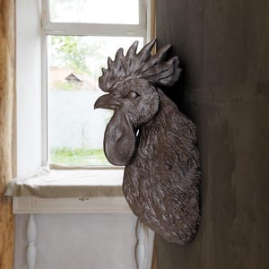 Puede incluir: Una escultura de cabeza de gallo marrón montada en una pared. La escultura está hecha de un material texturizado que se asemeja a plumas. La cabeza del gallo está girada hacia un lado y su pico está abierto.