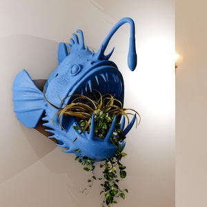 Puede incluir: Un macetero de pez pescador azul con una planta que crece de su boca. El macetero está montado en una pared.