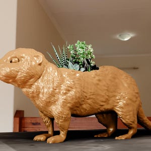 May include: A golden brown ceramic planter shaped like an otter with a variety of green succulents growing inside.