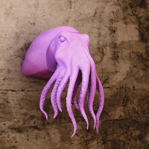 Könnte beinhalten: Eine lavendelfarbene Oktopus-Wandskulptur mit detaillierten Tentakeln und einer strukturierten Oberfläche. Die Skulptur ist an einer gesprenkelten braunen und beigen Wand angebracht und erzeugt einen auffälligen Kontrast.