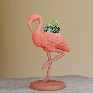 May include: A pink flamingo planter with a single leg and a base. The planter is filled with green succulents.