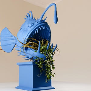 May include: A blue anglerfish planter with a green plant growing out of its mouth. The planter is on a blue pedestal.