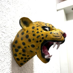 Puede incluir: Escultura mural de una cabeza de jaguar. La escultura está pintada con un patrón de manchas amarillas y negras, con una expresión de enfado, la boca abierta y dientes blancos visibles. El interior de la boca es rosa.