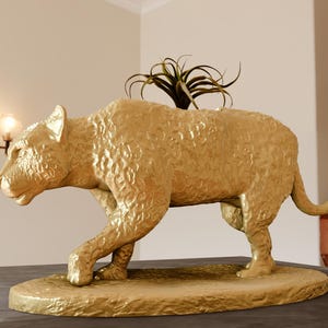Könnte beinhalten: Ein goldfarbener Tier-Pflanztopf in Form eines hockenden Leoparden. Der Topf hat eine strukturierte Oberfläche und beherbergt eine kleine grüne Pflanze.
