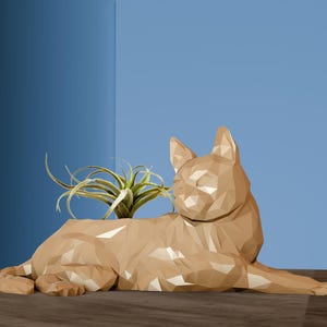 May include: A geometric, gold-coloured cat planter with a green air plant inside. The planter is made of a smooth, textured material and is in a relaxed, lying down pose.