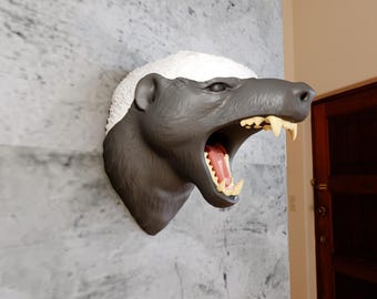 May include: A detailed wall-mounted sculpture of a honey badger head. The sculpture features a dark gray face with an open mouth revealing a pink interior and yellow teeth. The top of the head is white and textured.