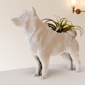 May include: A white ceramic dog planter with a textured surface, holding a green plant. The dog is standing on a white surface. The planter is a decorative item for home decor.