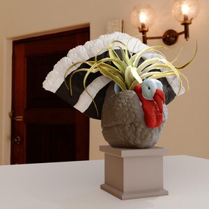 May include: A ceramic turkey planter with a white and black feathered tail, holding a green air plant. The planter is on a grey pedestal.