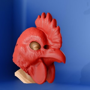 May include: A bright red chicken mask with a detailed comb, beak, and wattle. The mask is made of a smooth material and is set against a blue background. The eye holes are visible, and the mask is mounted on a light-colored base.