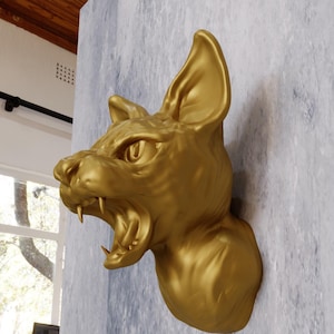May include: A gold-colored, snarling cat head sculpture with an open mouth and sharp teeth. The sculpture is mounted on a wall.