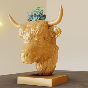 Op de afbeelding: Gouden keramische sculptuur van een Highland-koeienkop met een kleine vetplant die er bovenop groeit. De sculptuur staat op een gouden voet.