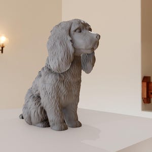 Peut inclure: Une sculpture grise d'un chien aux longues oreilles tombantes, assis sur une surface blanche. La statue présente une fourrure texturée et une expression paisible. L'arrière-plan comprend un mur avec une applique et une poutre en bois.