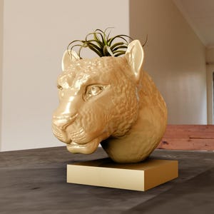 Könnte beinhalten: Goldfarbener Keramik-Pflanztopf in Form eines Leopardenkopfes mit einer kleinen grünen Pflanze, die aus der Oberseite wächst.