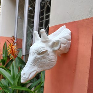 May include: White ceramic giraffe head wall decor. The giraffe head is mounted on a wall with a pink and white background. The giraffe has a detailed face and a textured pattern on its neck.