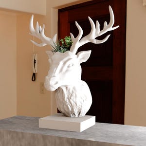 May include: White ceramic deer head sculpture with antlers, used as a planter with green plants growing out of the top. The sculpture is on a white pedestal and a grey surface.
